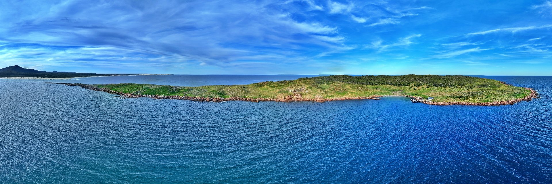 Aerial panorama of Gabo Island from the sea, with a rocky cove, green headland and the East Gippsland coast beyond.