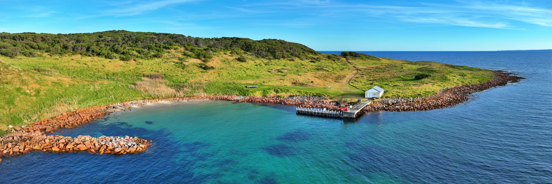 Aerial panorama of Gabo Island harbour, with a granite breakwater, jetty, white shed and turquoise water in the cove.