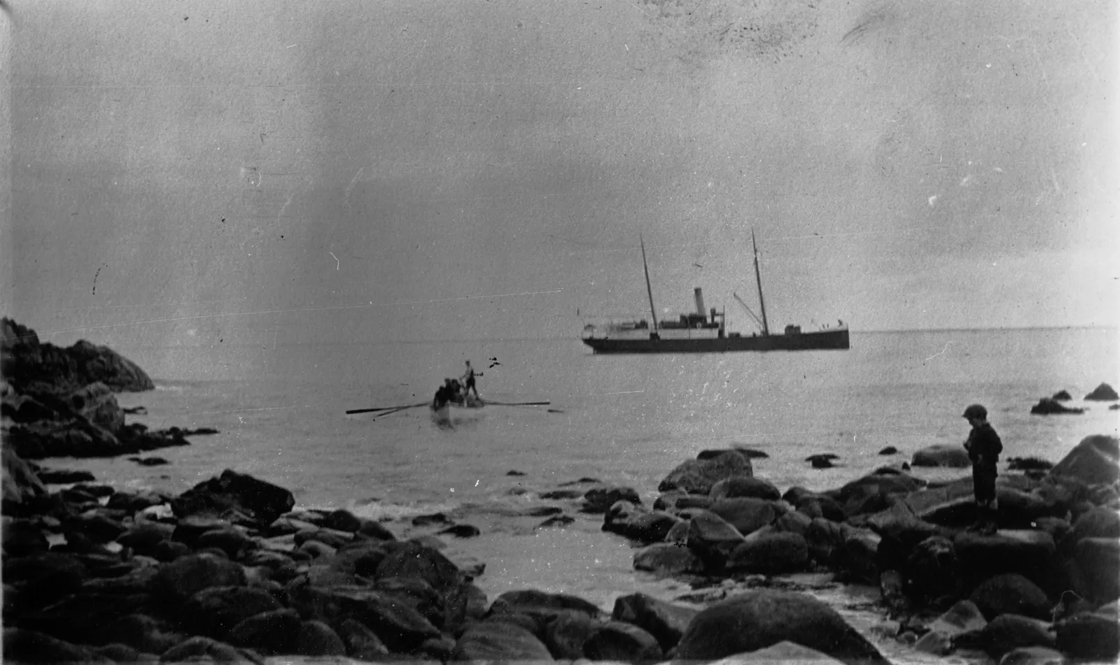 Black and white photograph of the two-masted steamer Lady Loch lying offshore while a rowing boat loaded with stores heads for a rocky beach.